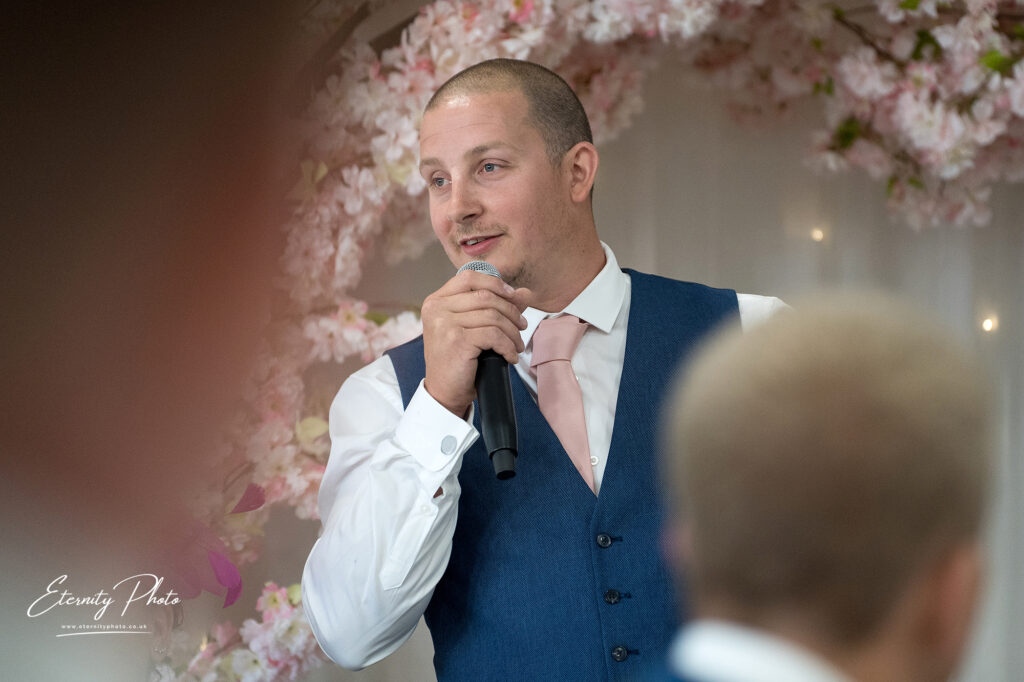 A man in a vest holding a microphone and speaking at an event with a floral backdrop.