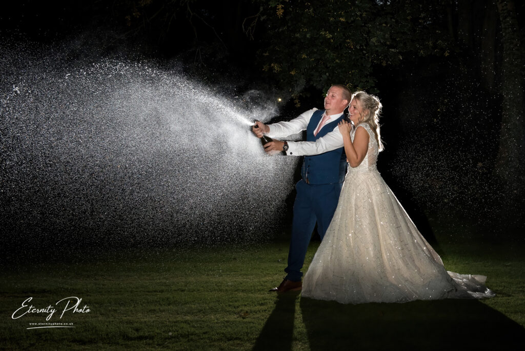 Bride and groom spray champagne at night with a backlight highlighting the spray.