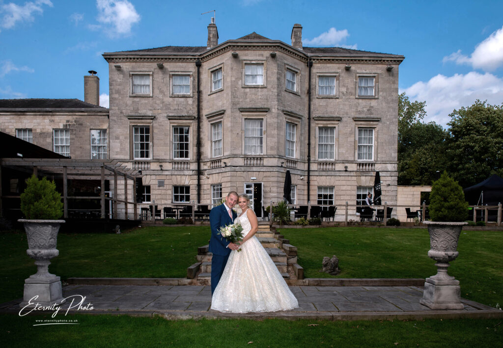 Bride and groom posing for a photo in front of an elegant manor house with a clear blue sky overhead.