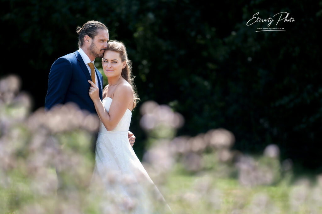 A bride and groom embracing outdoors, with the focus on them behind a blur of flowers.