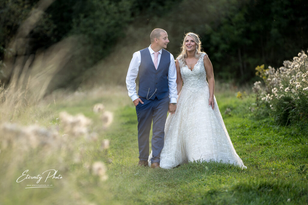 A bride and groom holding hands and smiling at each other in a sunlit meadow.
