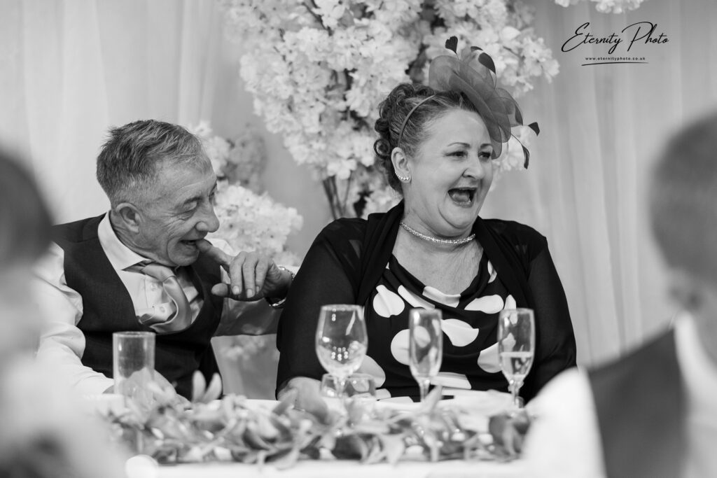 A woman laughs heartily while sitting at a table during a social event, with a man to her left smiling in her direction.