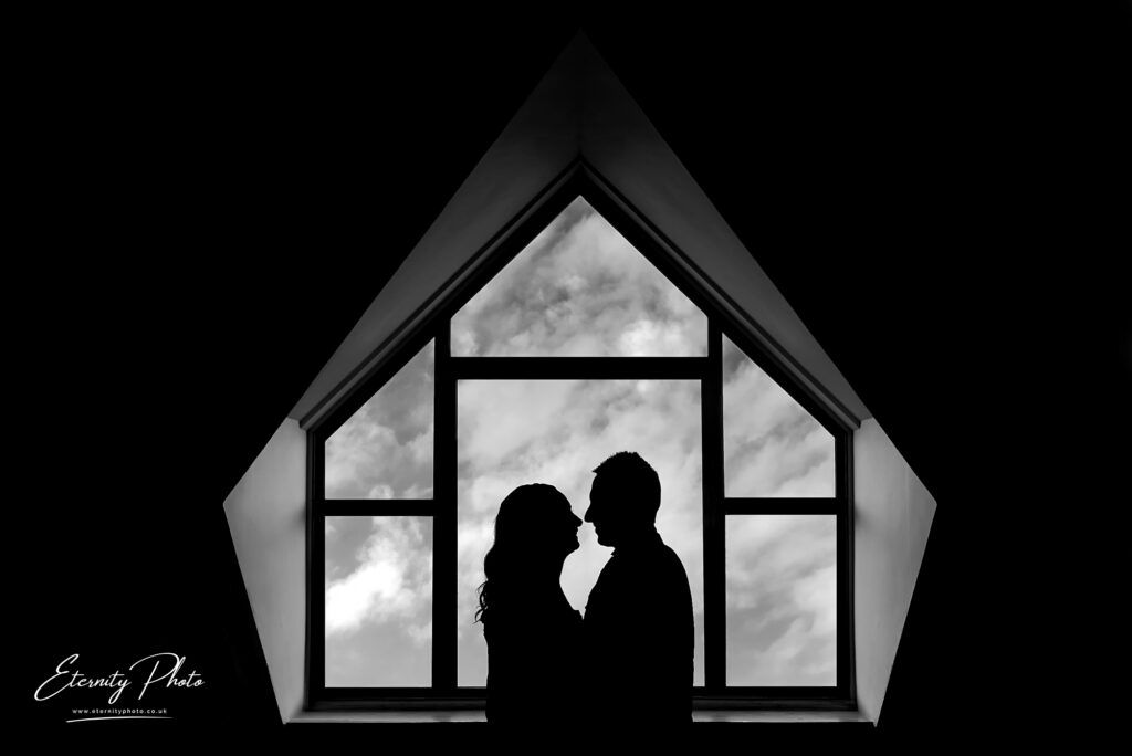 Silhouettes of a couple against a cloudy sky viewed through a triangular window.