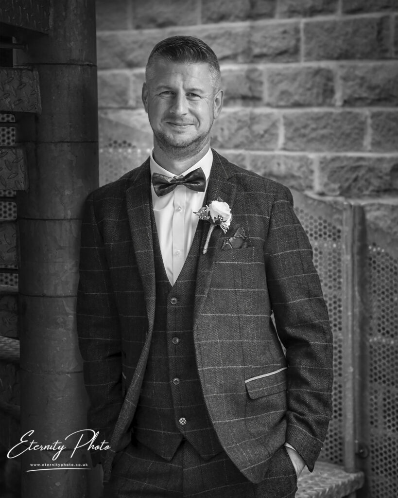 A monochrome portrait of a well-dressed man with a bow tie and a boutonniere, standing confidently against a textured backdrop.