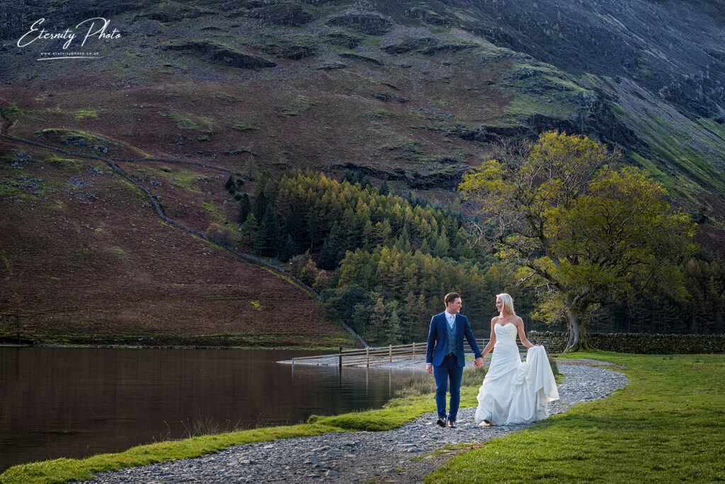 A couple in wedding attire holding hands by a serene lake with mountains in the background.