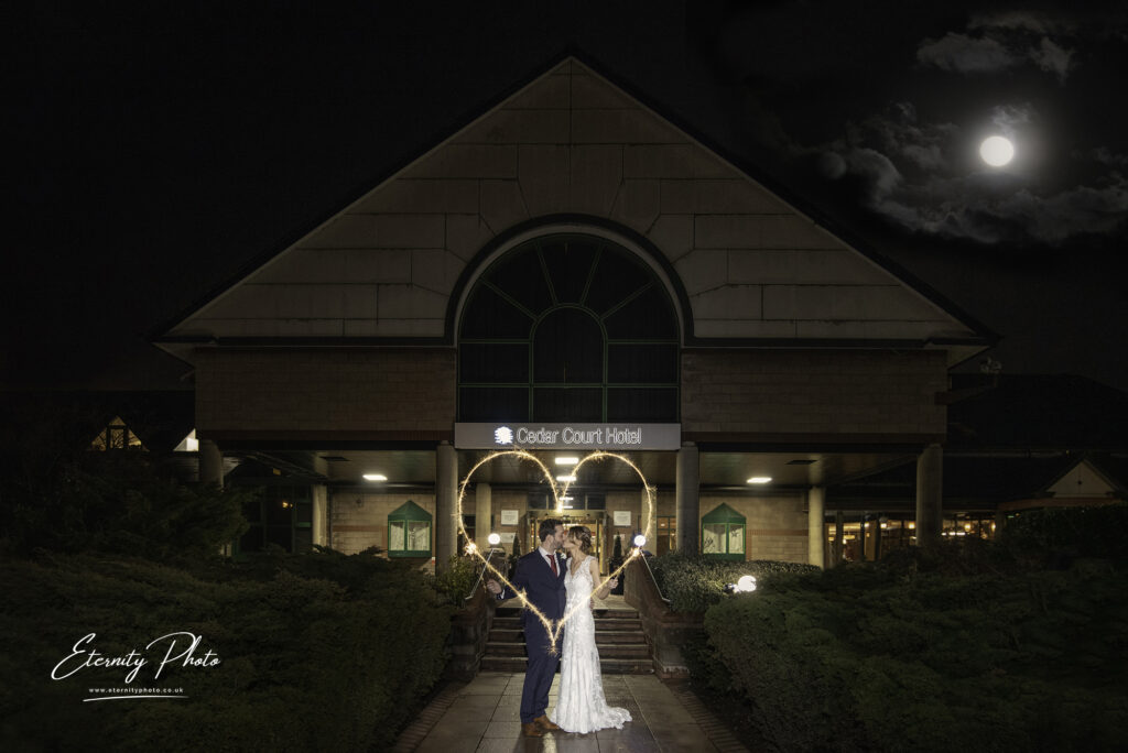 A couple in wedding attire holding hands in front of a hotel entrance under a night sky with a full moon.