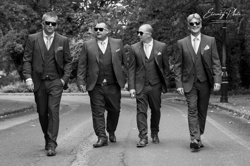 Four men in suits and sunglasses walking confidently down a road.