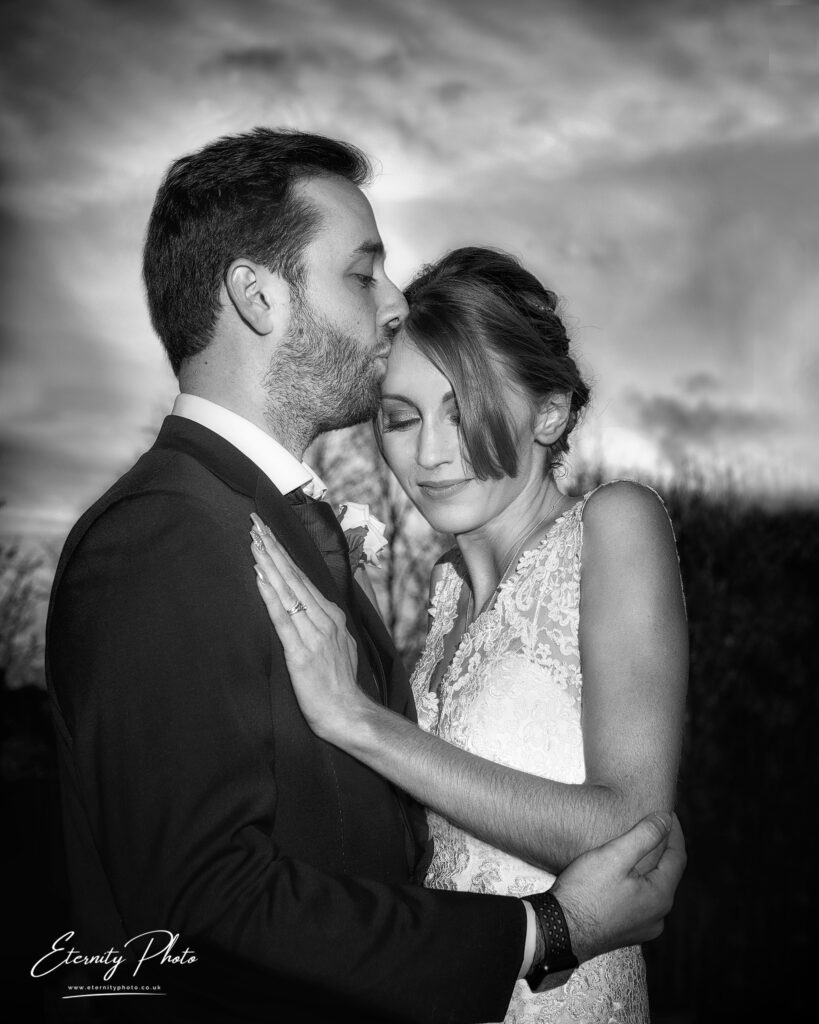 A monochrome image of a woman in a lace wedding dress and a man in a suit embracing and touching foreheads under a dramatic sky.