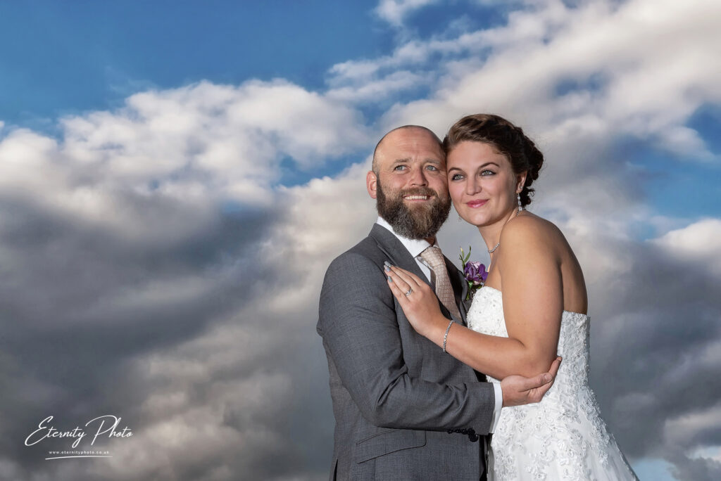 A couple dressed in wedding attire embracing against a backdrop of dramatic clouds.
