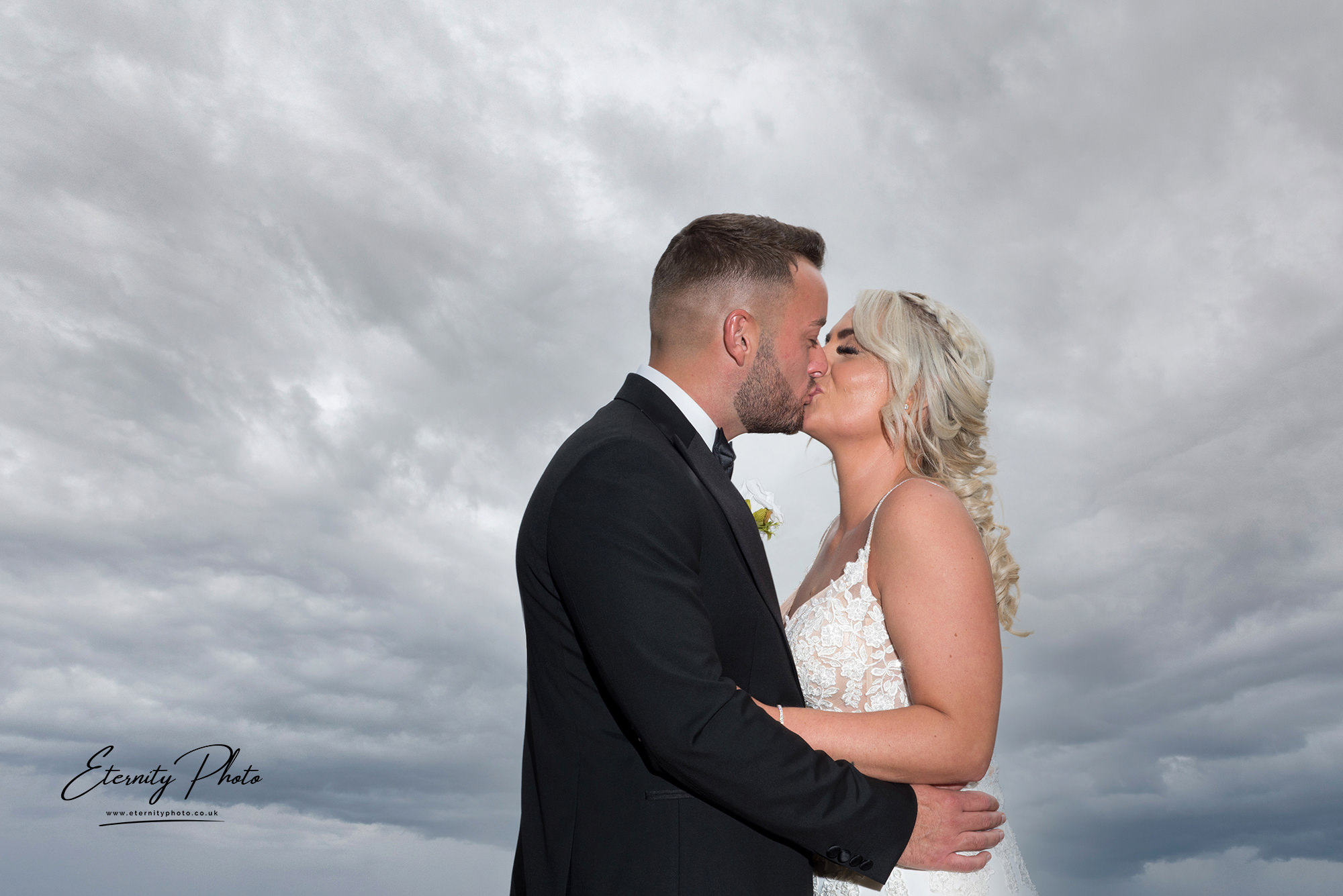 Couple kissing under dramatic clouds on wedding day.