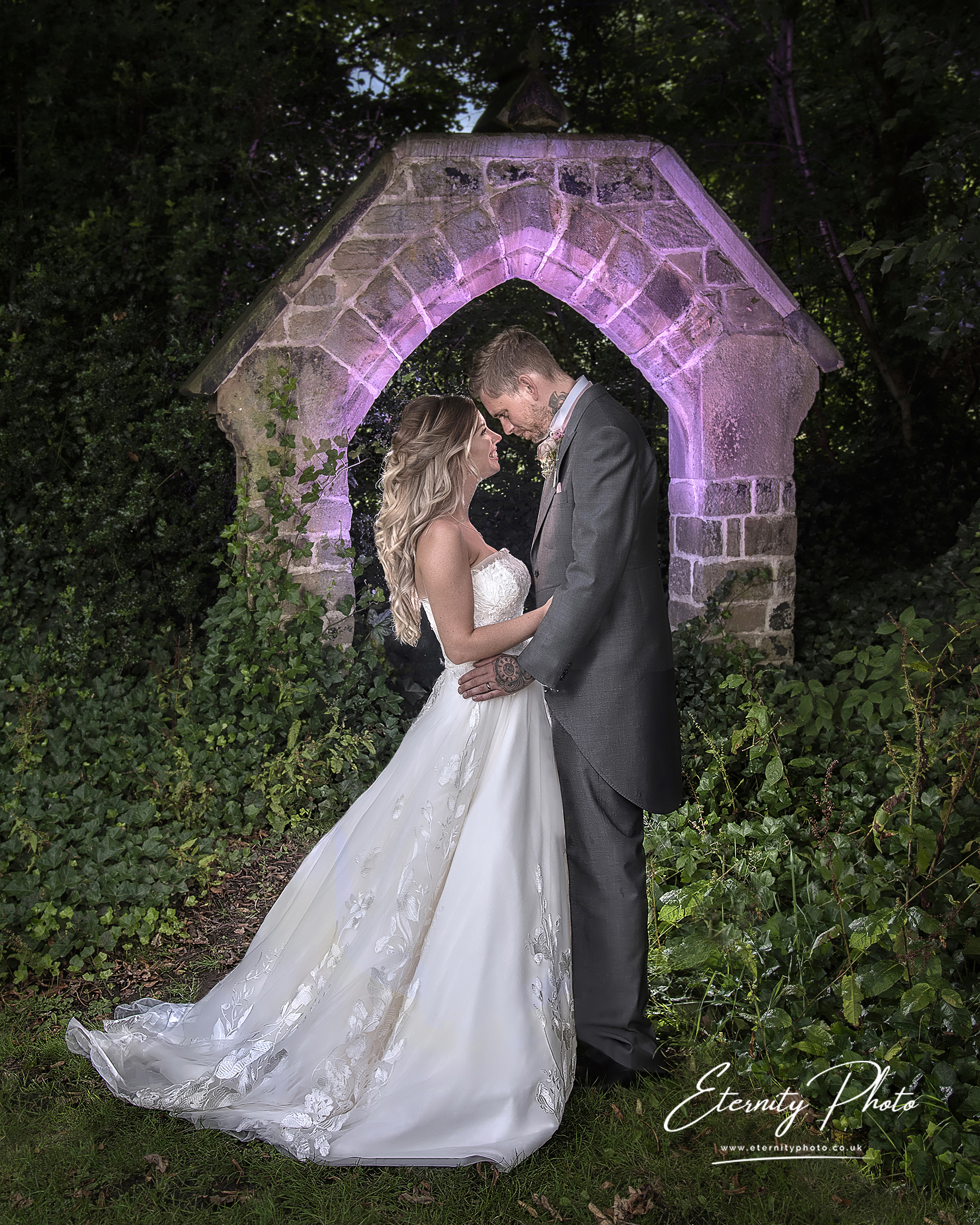 Bride and groom embracing under stone archway outdoors.