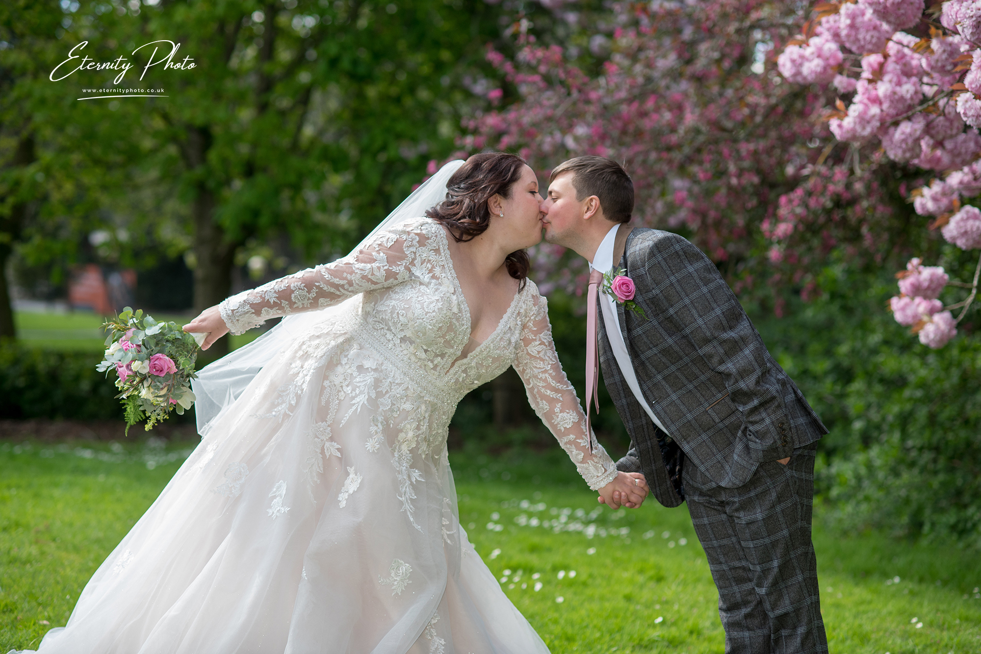 Bride and groom kissing by blossoming trees at park.