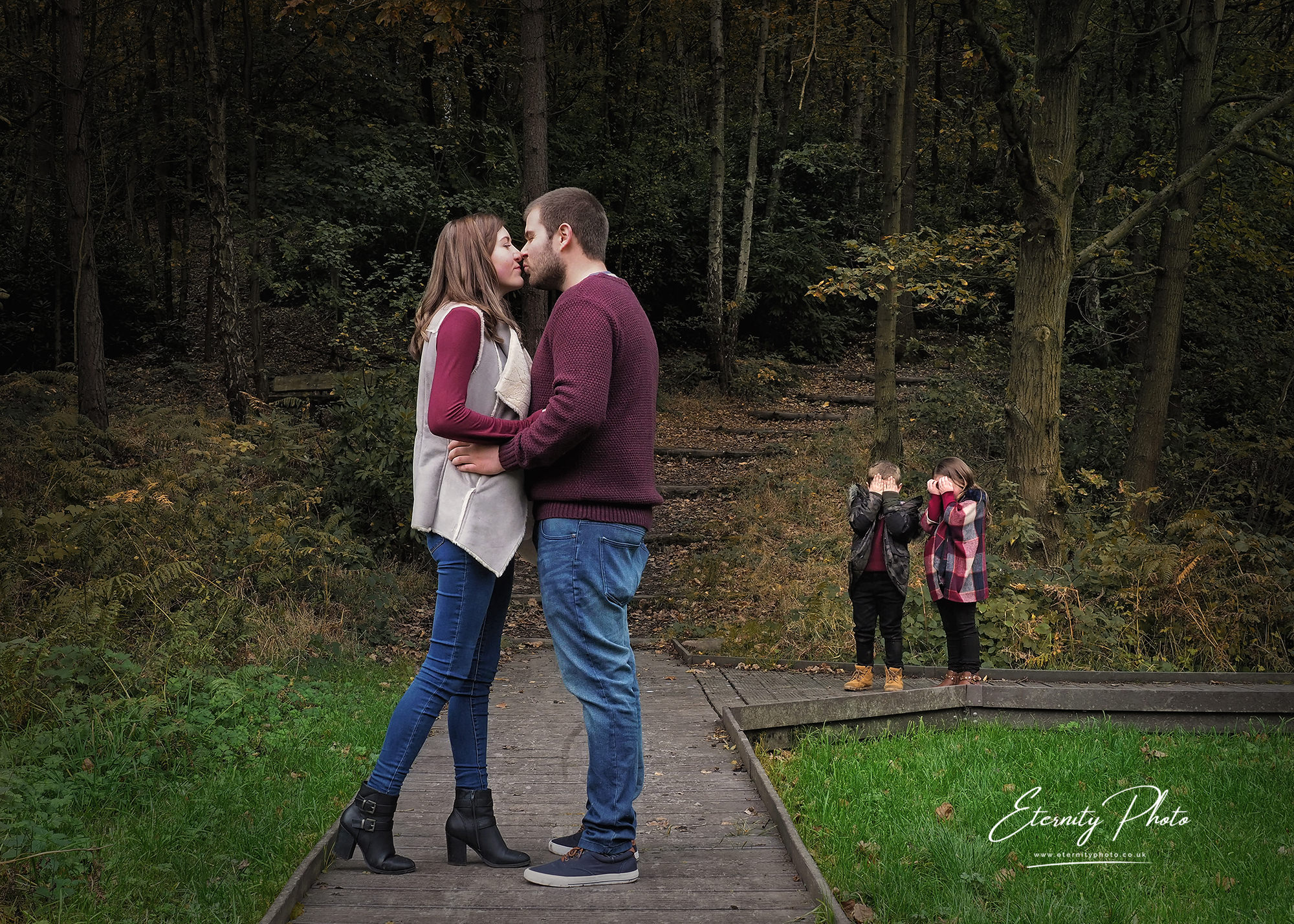 Couple kissing with children covering eyes in forest.