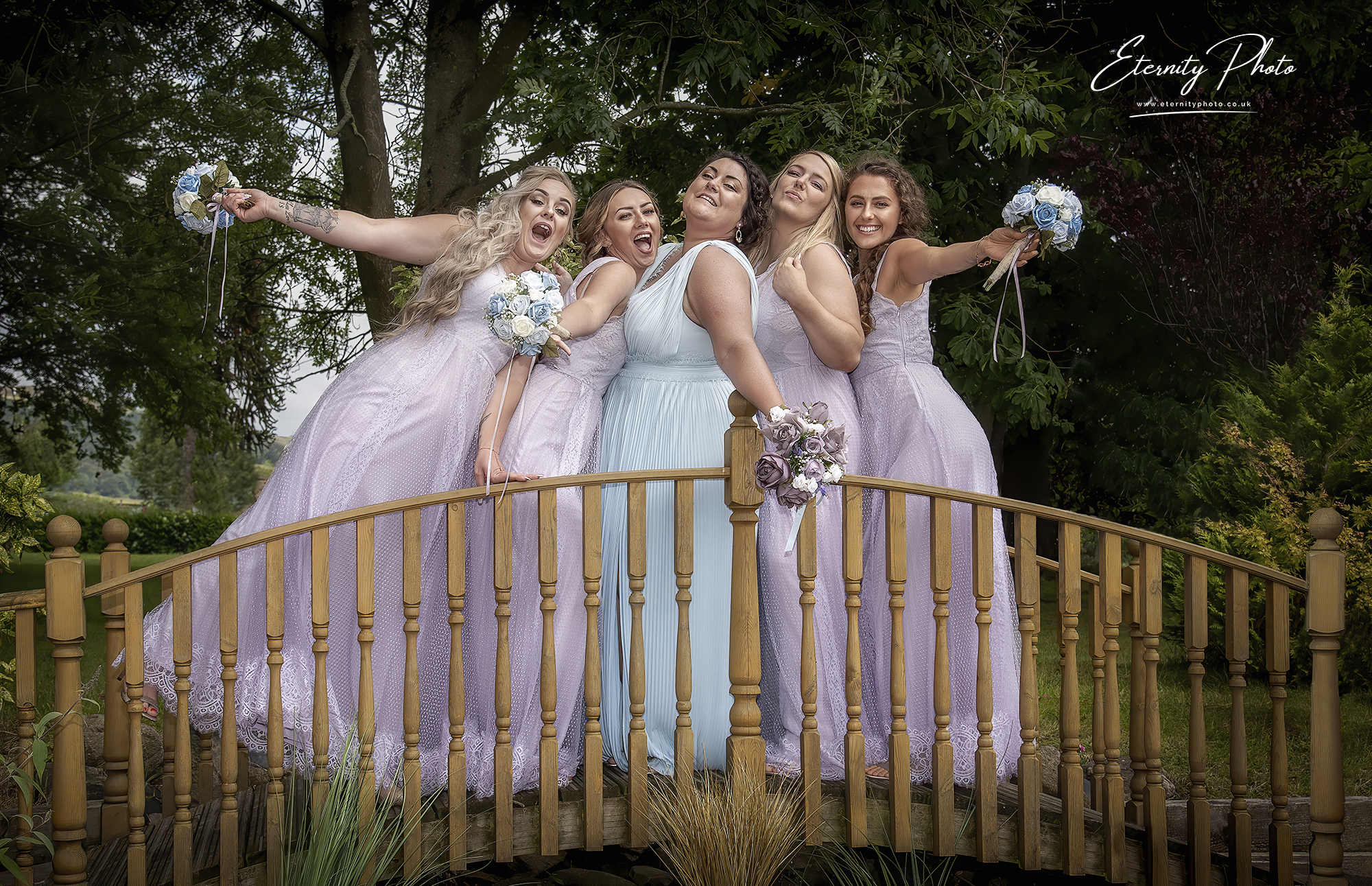 Bridesmaids smiling on bridge in lavender dresses with bouquets.