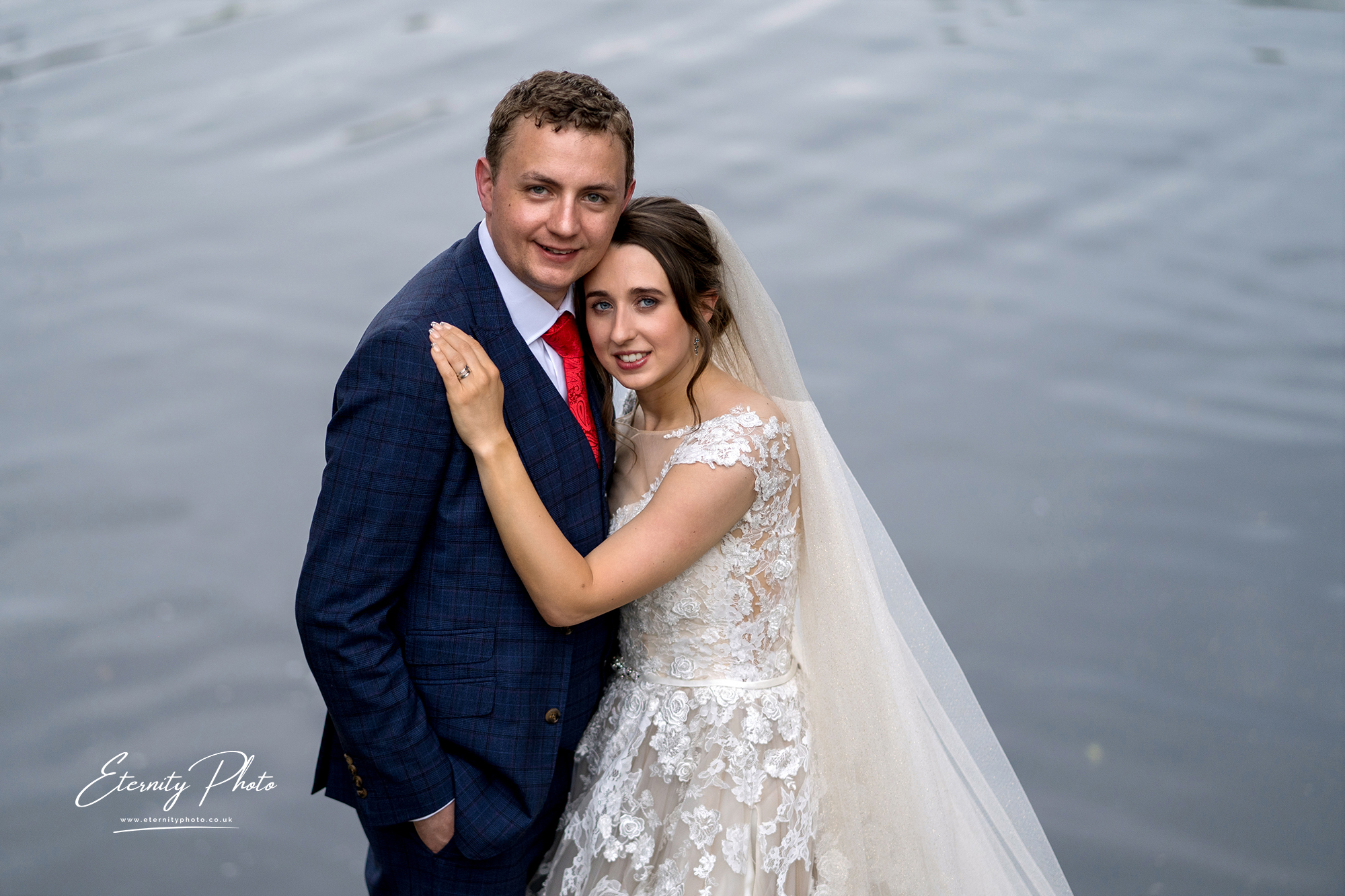 Bride and groom posing by water, wedding day photograph.